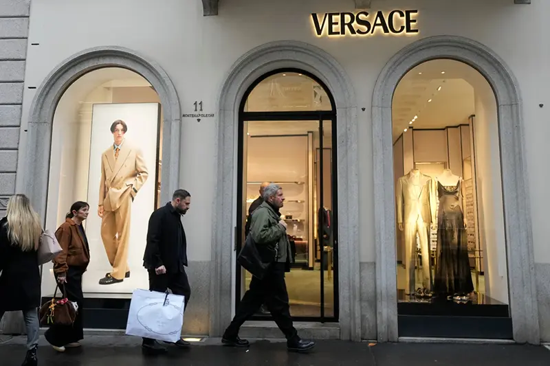 A man walks with a Prada shopping bag in front of a Versace shop, at the Montenapoleone luxury fashion street, in Milan, Italy, Tuesday, Dec. 2, 2025. (AP Photo/Luca Bruno)